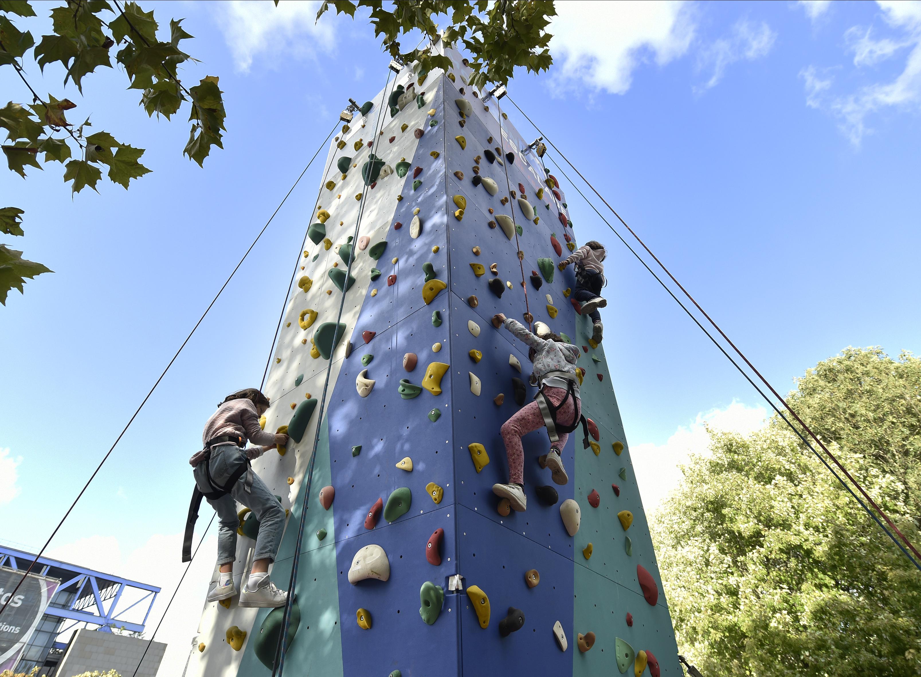 Enfants grimpant sur un mur d'escalade extérieur multicolore, sous un ciel bleu.
