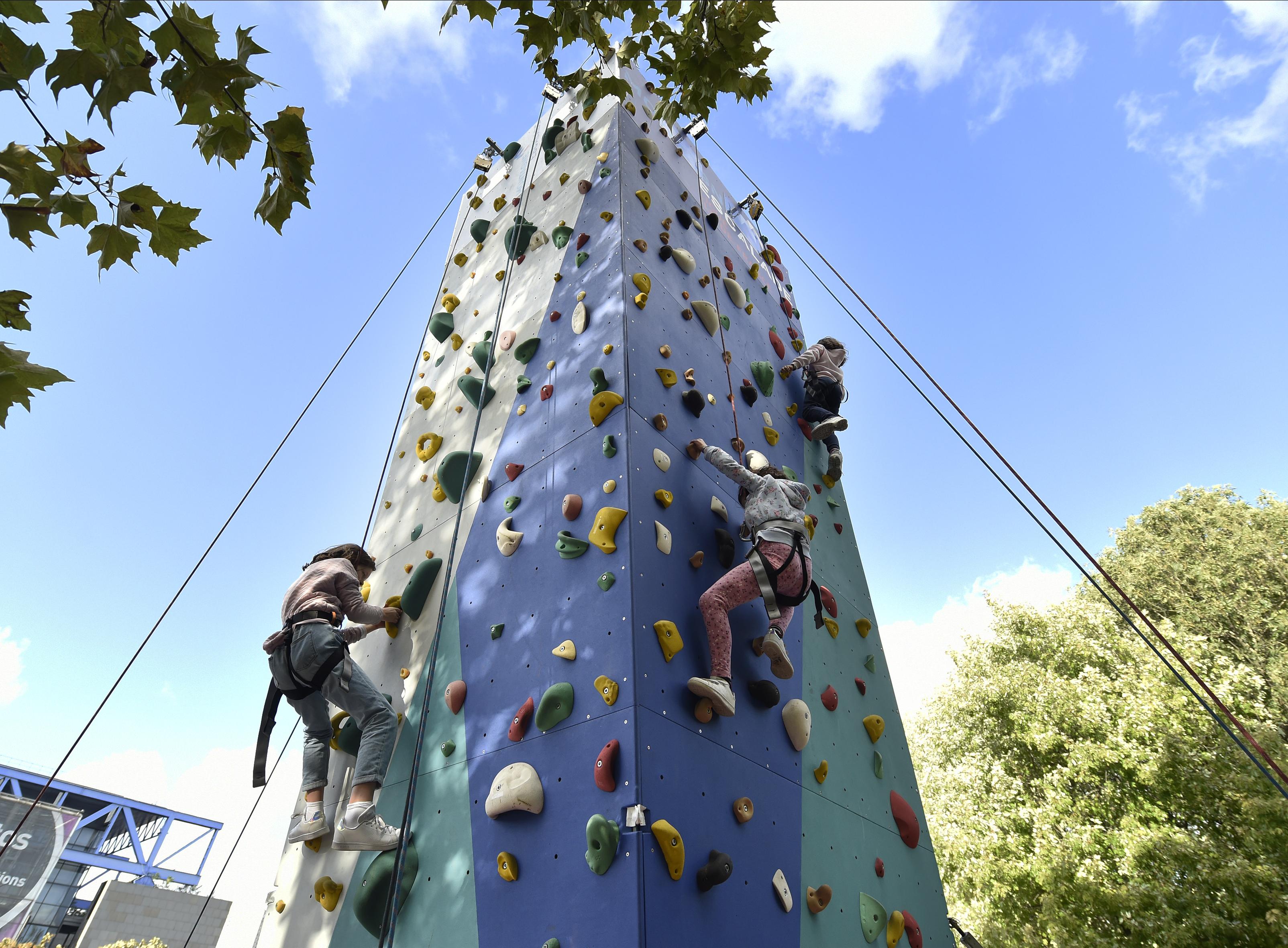 Enfants grimpant sur un mur d'escalade extérieur multicolore, sous un ciel bleu.