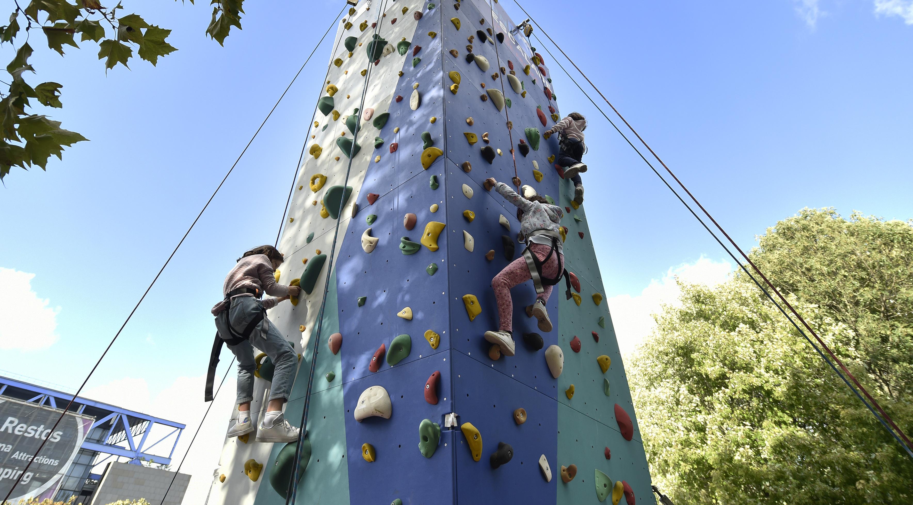 Enfants grimpant sur un mur d'escalade extérieur multicolore, sous un ciel bleu.
